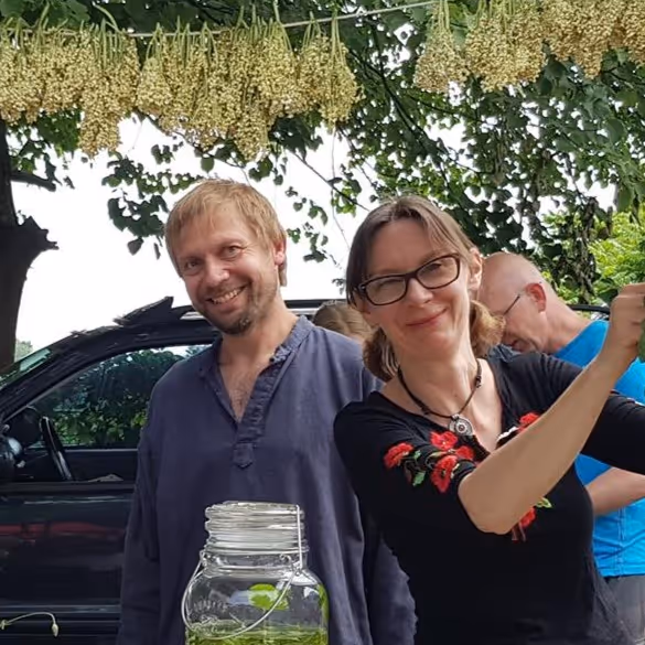 Para uśmiechających się osób na zewnątrz, obok nich słoik z zielonym napojem, z tle samochód i wiszące kwiaty nad nimi.