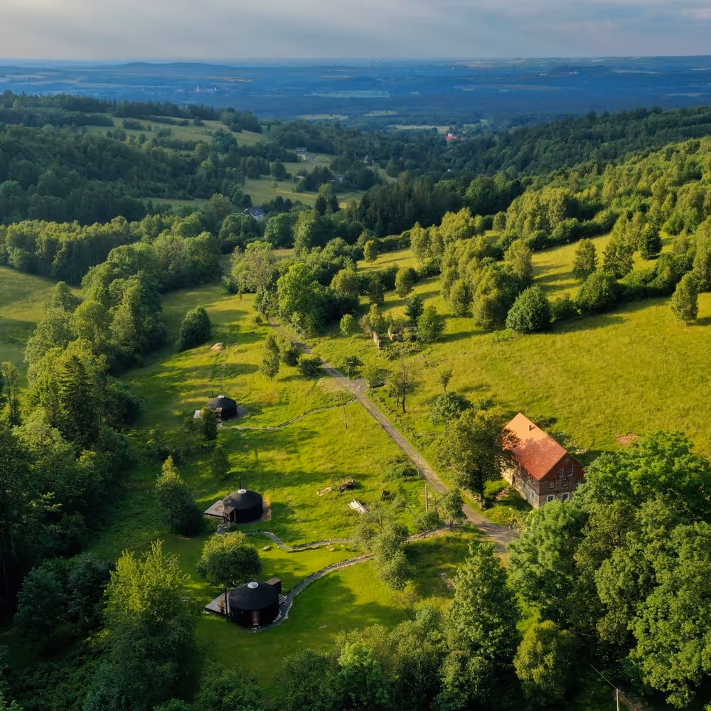 Widok z lotu ptaka na zielone wzgórza z trzema okrągłymi, czarnymi domkami i tradycyjnym budynkiem z czerwonym dachem po prawej stronie.