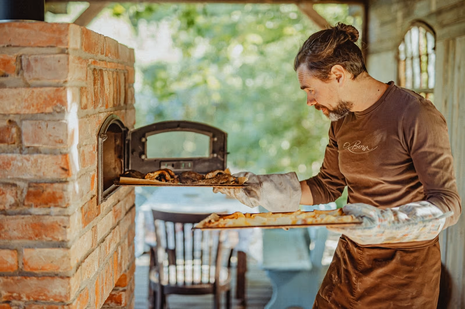 Mężczyzna w brązowej koszulce wkłada pizzę do pieca chlebowego z cegły na zewnątrz.