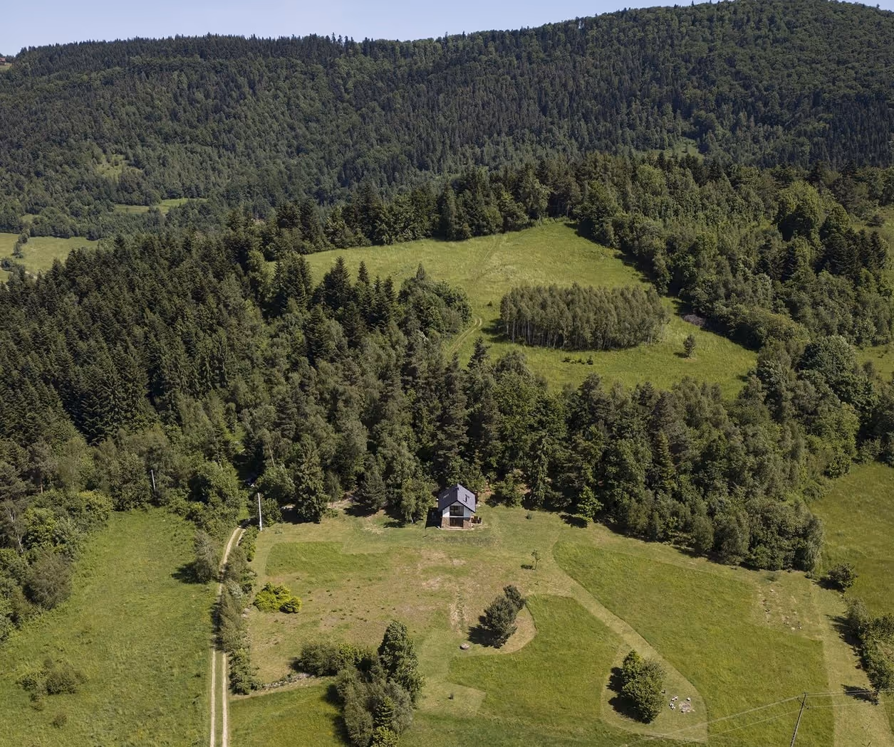 Dom położony na polanie otoczonej gęstym lasem na tle zalesionych wzgórz.