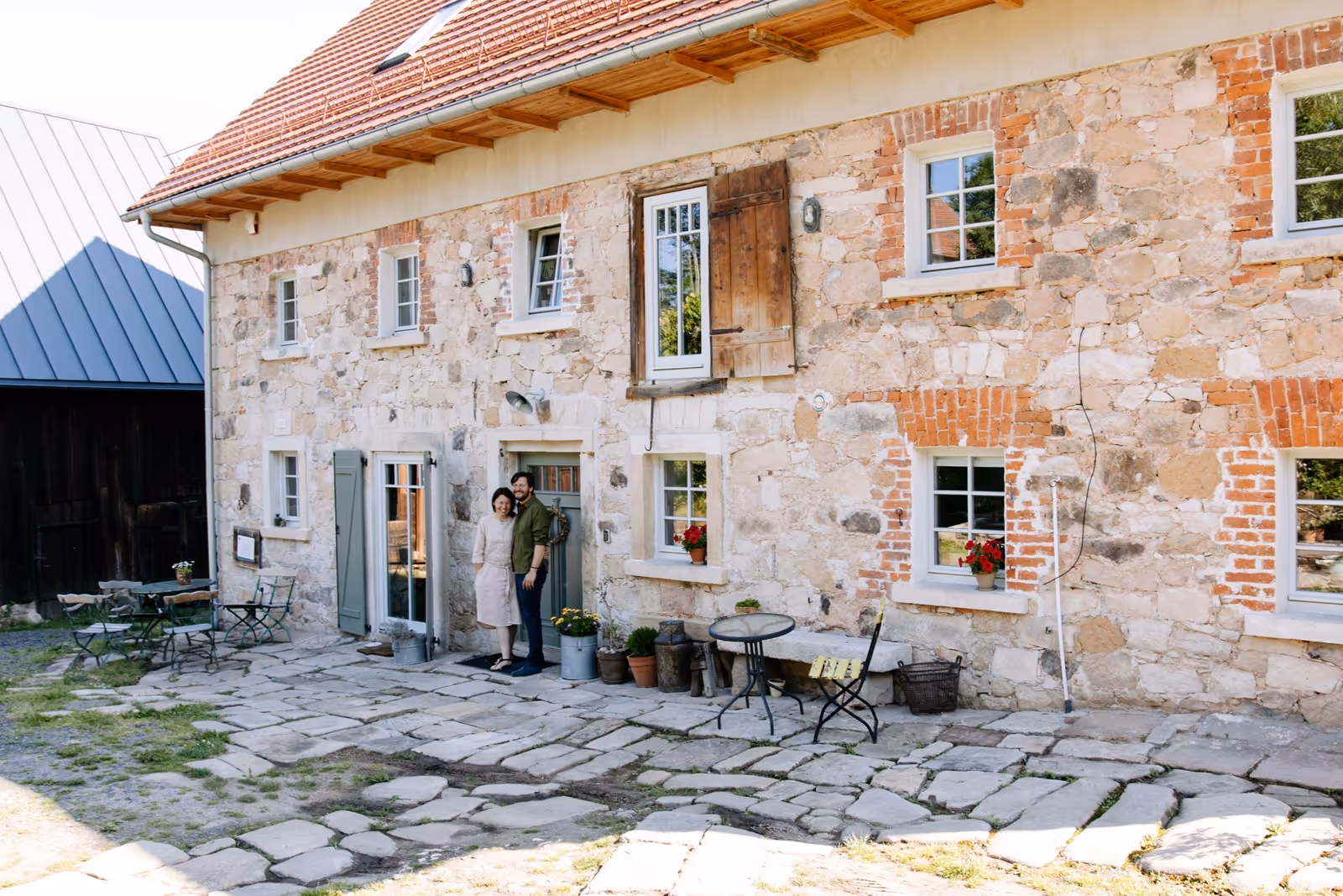 Para stojąca przy kamiennym domu z ceglanymi ozdobami i zielonymi drzwiami na patio z kamienną posadzką.