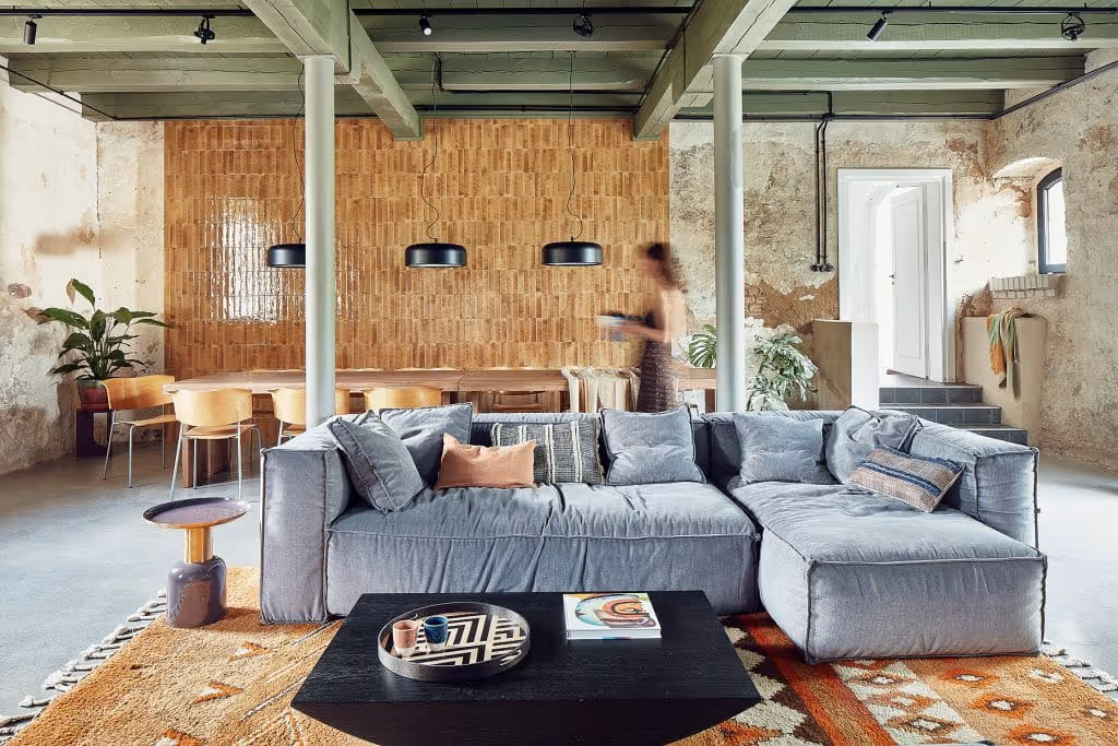 Przestronny salon w stylu loft z szarą narożną sofą, czarnym stolikiem kawowym, dywanem w ciepłych barwach oraz dużym drewnianym stołem i krzesłami w tle.
