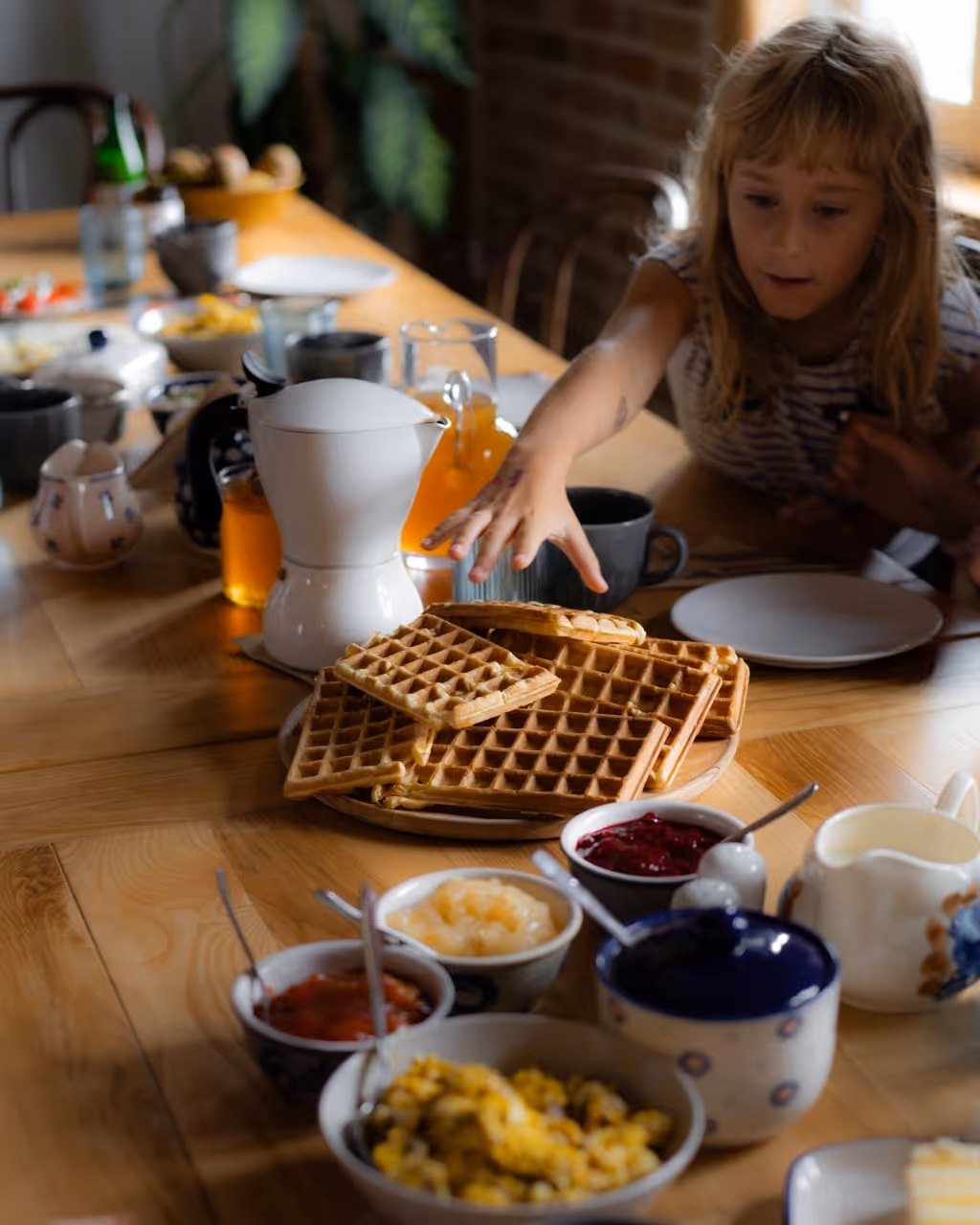 Dziewczynka sięga po stos gofrownic na stole kuchennym z różnymi dodatkami do śniadania.