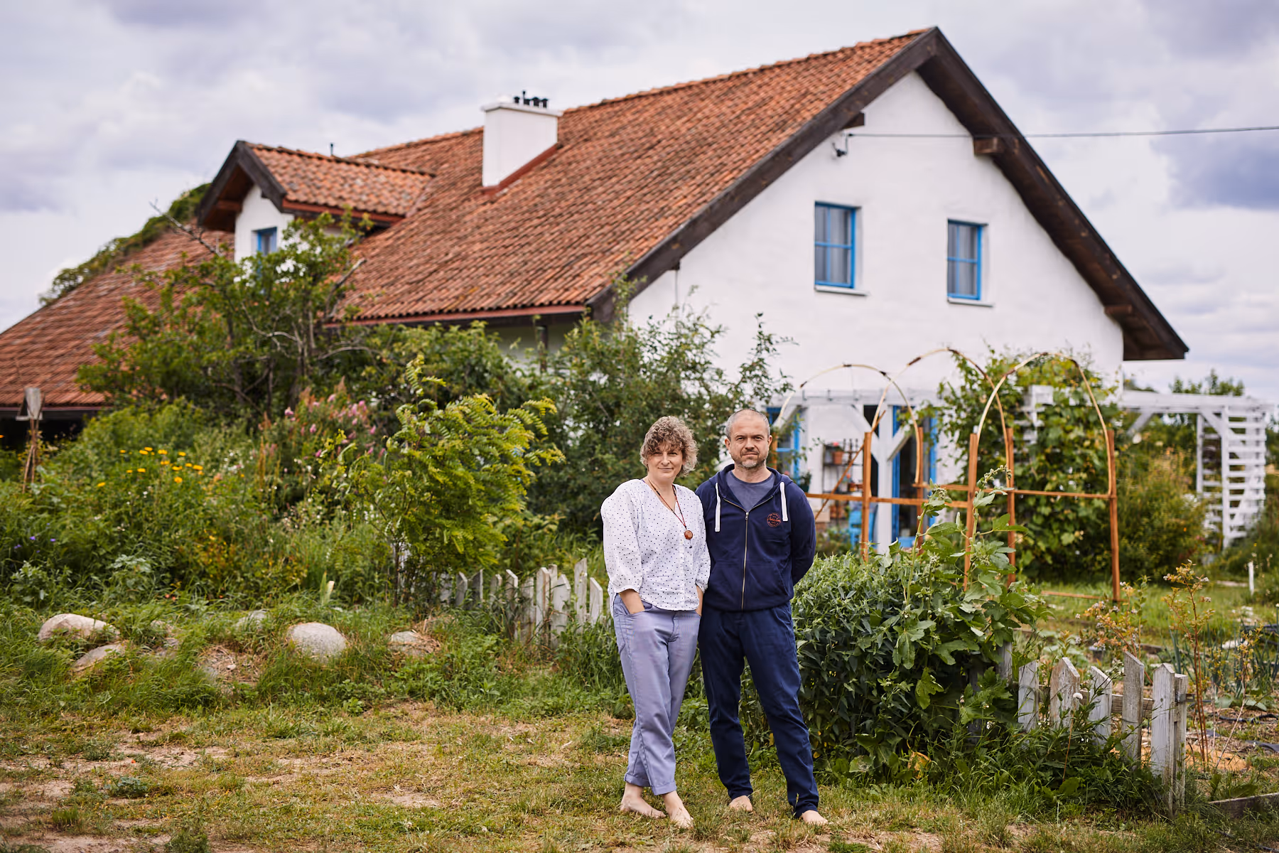 Para stojąca boso na trawie przed białym domem z czerwoną dachówką i ogrodem pełnym zieleni.