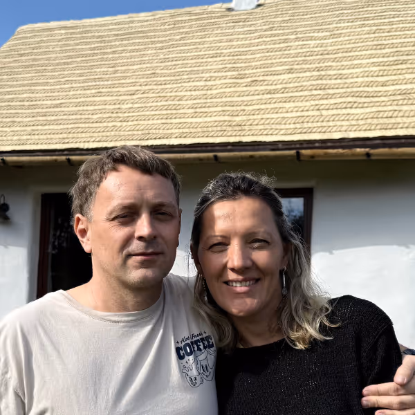 Para stojąca blisko siebie na tle domu z dachem słomianym, oboje uśmiechnięci.