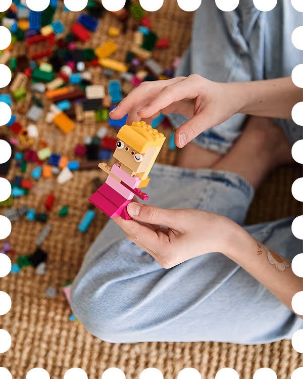 Dłonie osoby trzymającej kolorową figurkę z klocków LEGO, siedzącej na wykładzinie z rozrzuconymi klockami w tle.