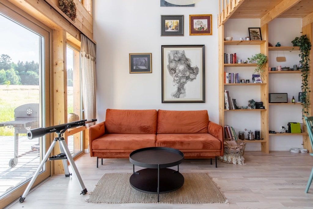 Nowoczesny salon z pomarańczową sofą, czarnym okrągłym stolikiem, teleskopem przy dużym oknie i drewnianymi półkami z książkami i roślinami.