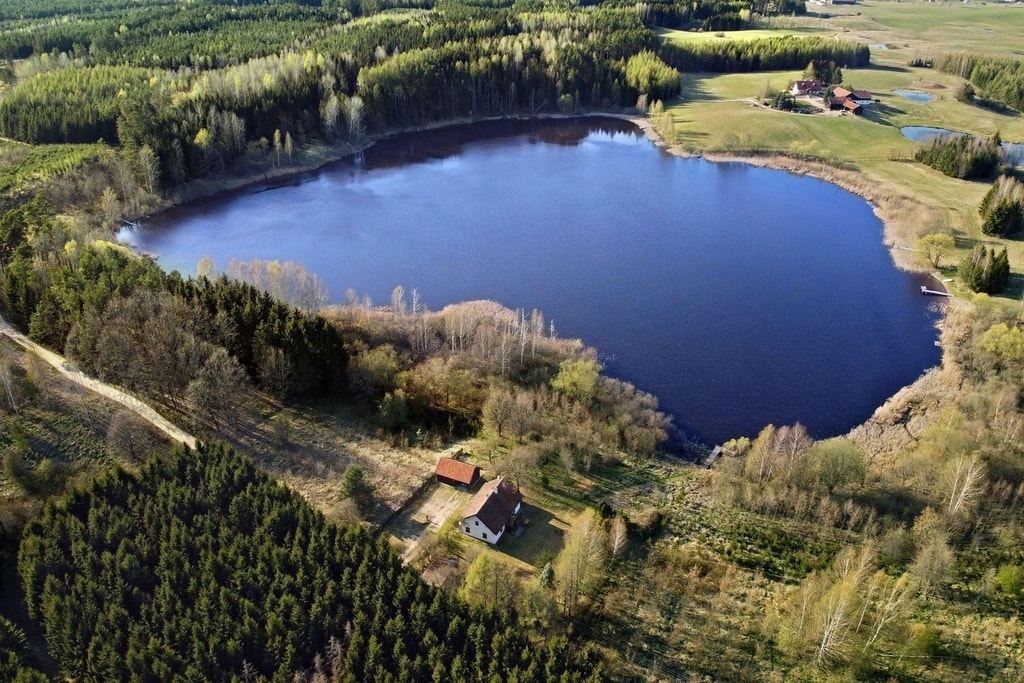 Jezioro otoczone lasem z małym domem i zabudowaniami w pobliżu, widok z lotu ptaka.